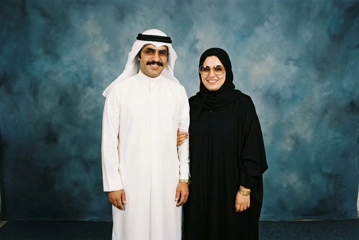 ستايل الثمانينات من FaceCast: زوجان بالغان في بورتريه استوديو، هو بثوب أبيض مكوي وغترة بيضاء وعقال أسود ونظارة أفياتور ذهبية وشنب وساعة ذهبية، وهي بعباية سوداء وشيلة سوداء تغطي الشعر ونظارات كبيرة وأساور ذهب، أمام خلفية استوديو زرقاء مرقطة بفلاش مباشر
