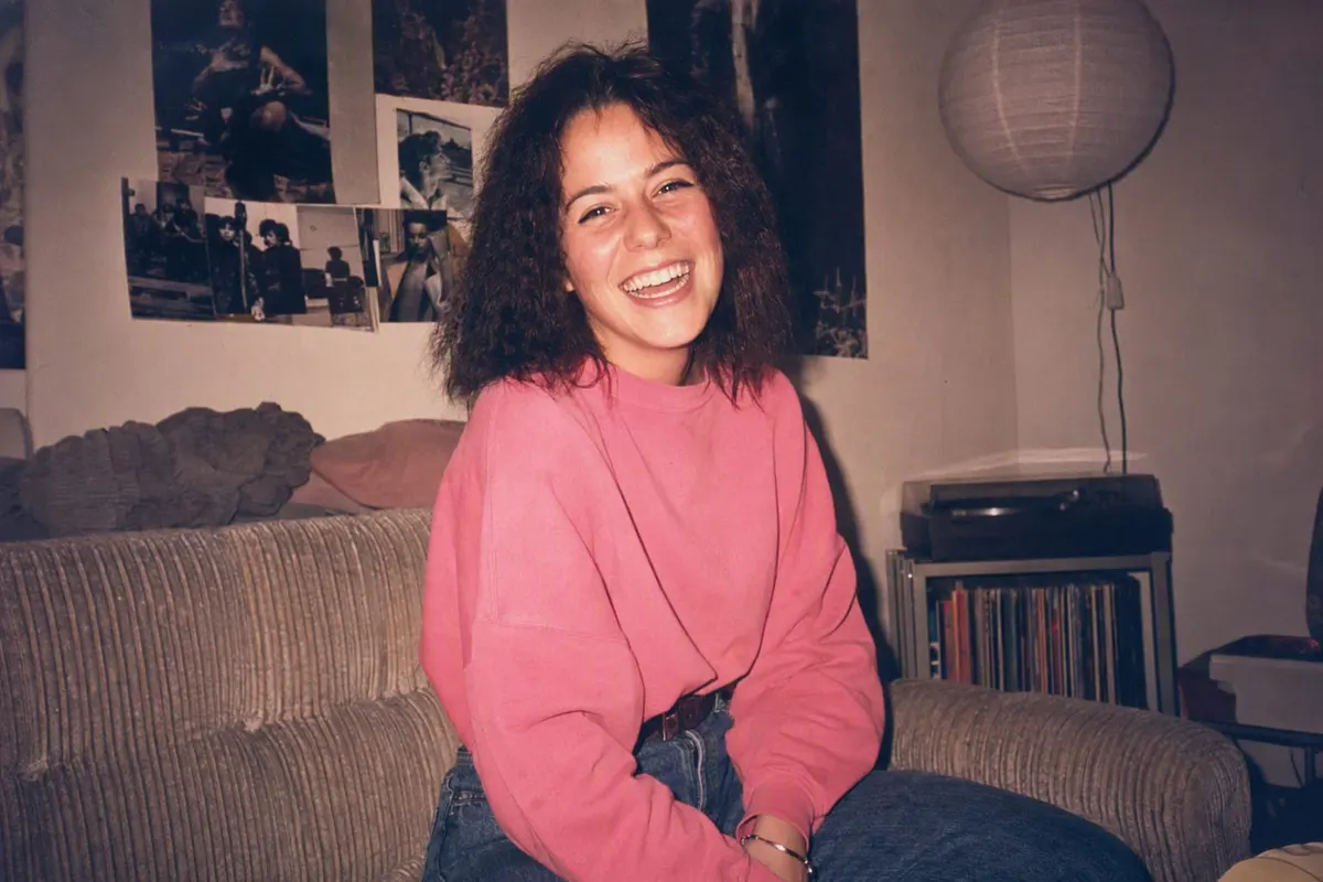 Junge Frau in einer WG 1986: gekreppte Haare, neonpinkes Sweatshirt, Plattenspieler und Plakatwand im Hintergrund, hartes Blitzlicht