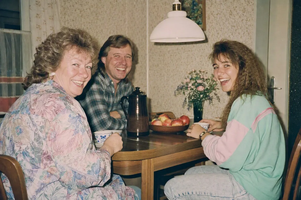 Familienschnappschuss am Küchentisch in einer westdeutschen Wohnung 1986: drei Erwachsene mit Dauerwelle und Vokuhila, gemusterte Tapete, hartes Blitzlicht