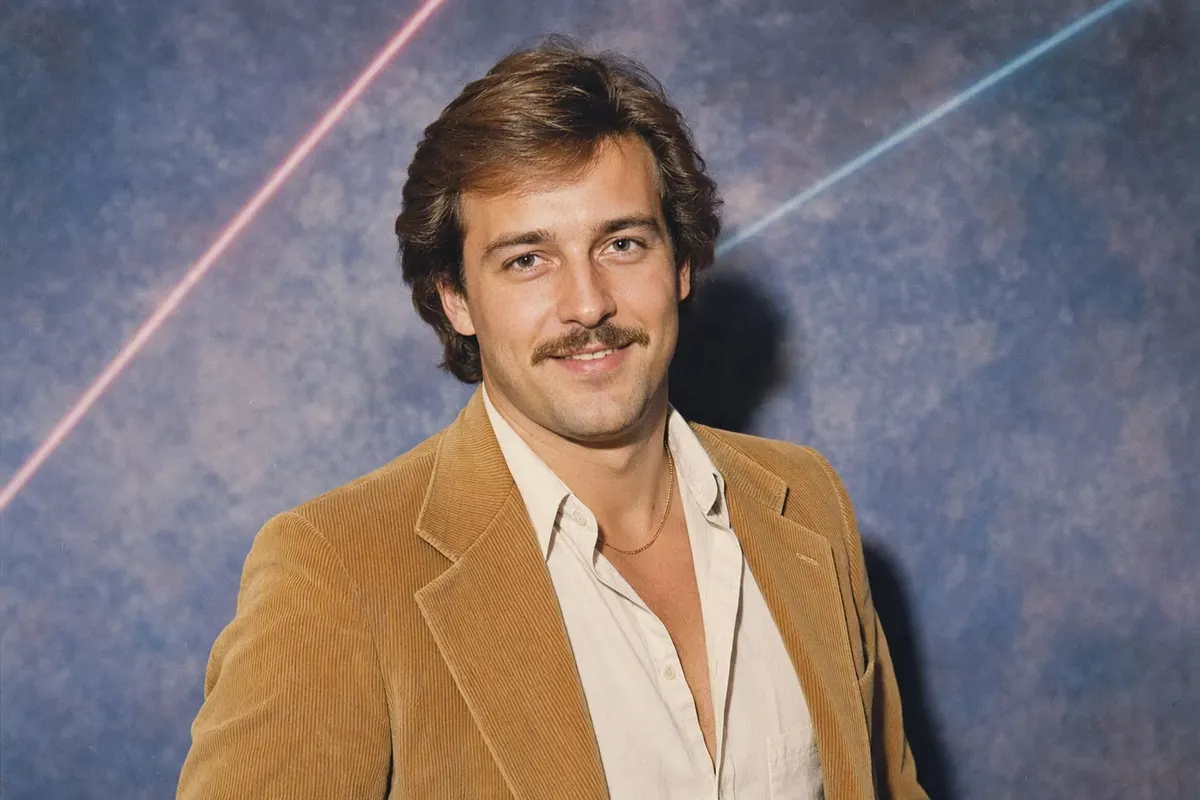 A 1986 studio portrait of a man in a tan corduroy blazer with wide lapels, lit by a hard direct flash