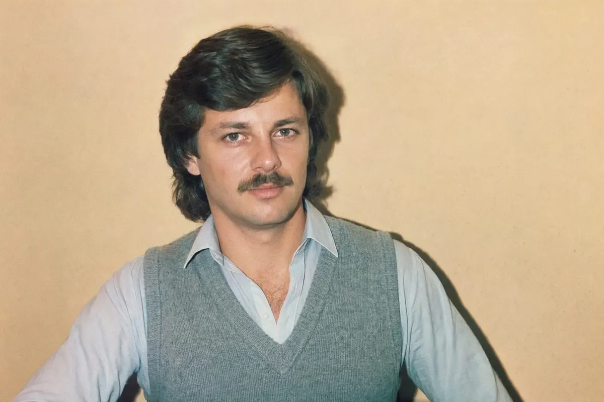 1986 snapshot of a man with feathered hair and a knit vest, lit by a hard direct on-camera flash against a warm beige wall