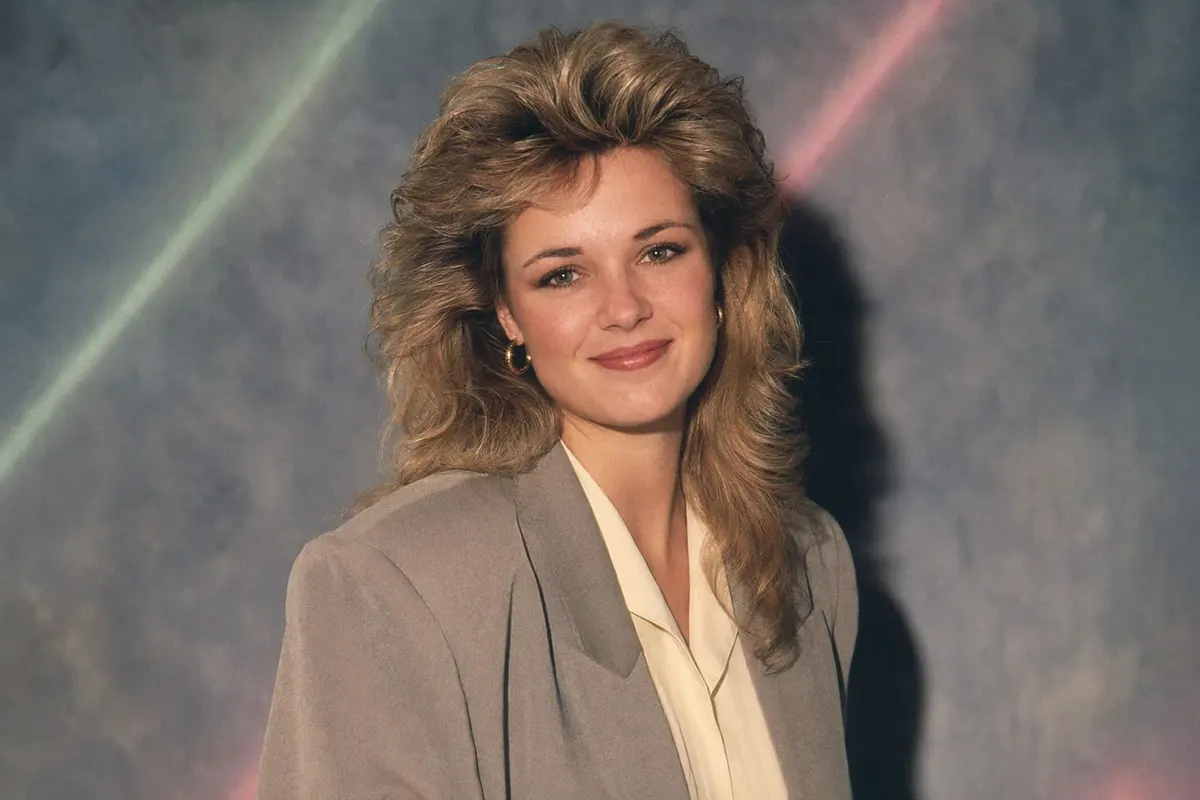 A 1986 studio portrait of a woman with big feathered hair and a grey blazer, lit by a hard direct flash against a mottled backdrop