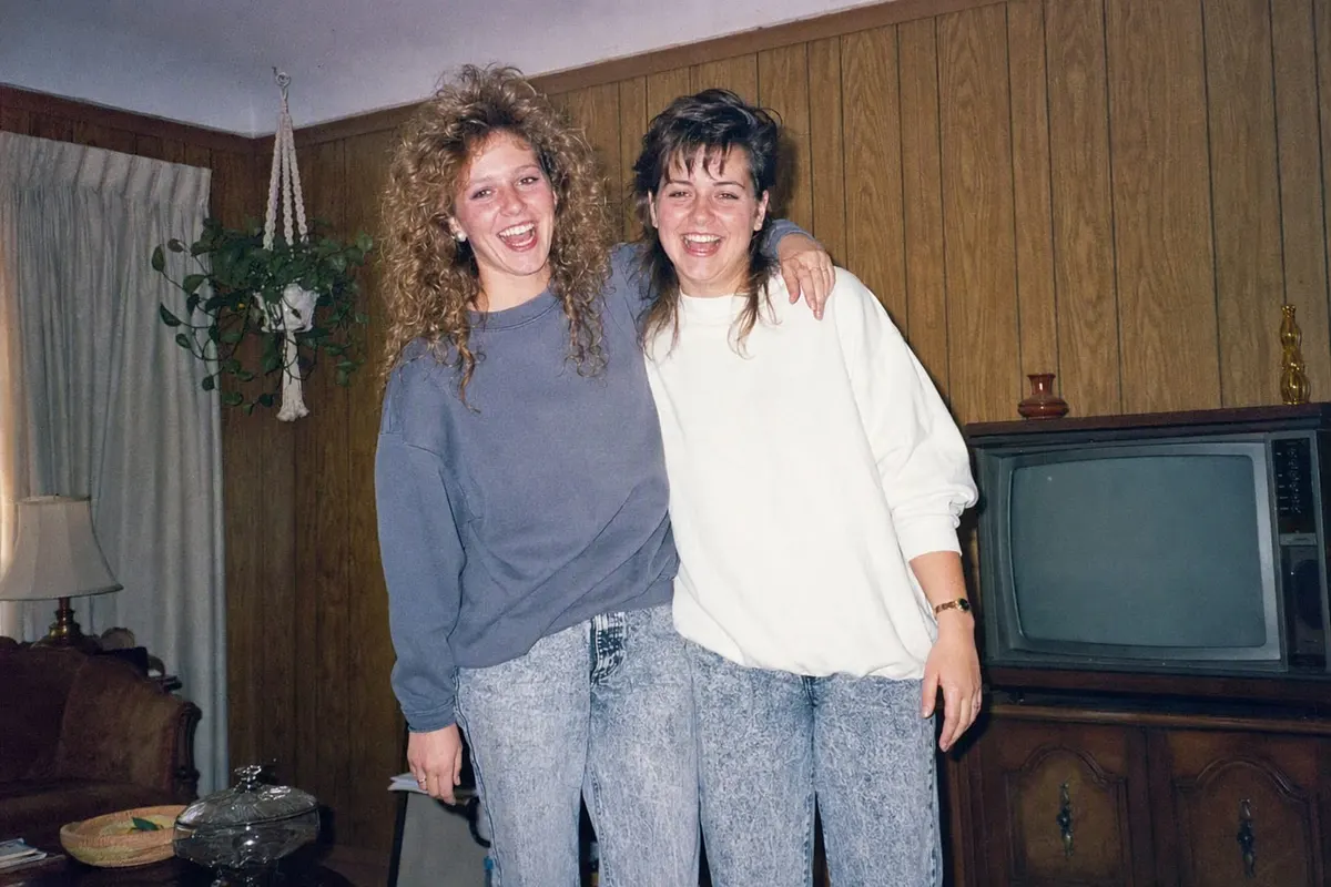 Two friends in a 1986 living room with permed hair, a mullet and acid-wash denim, lit by a hard on-camera flash