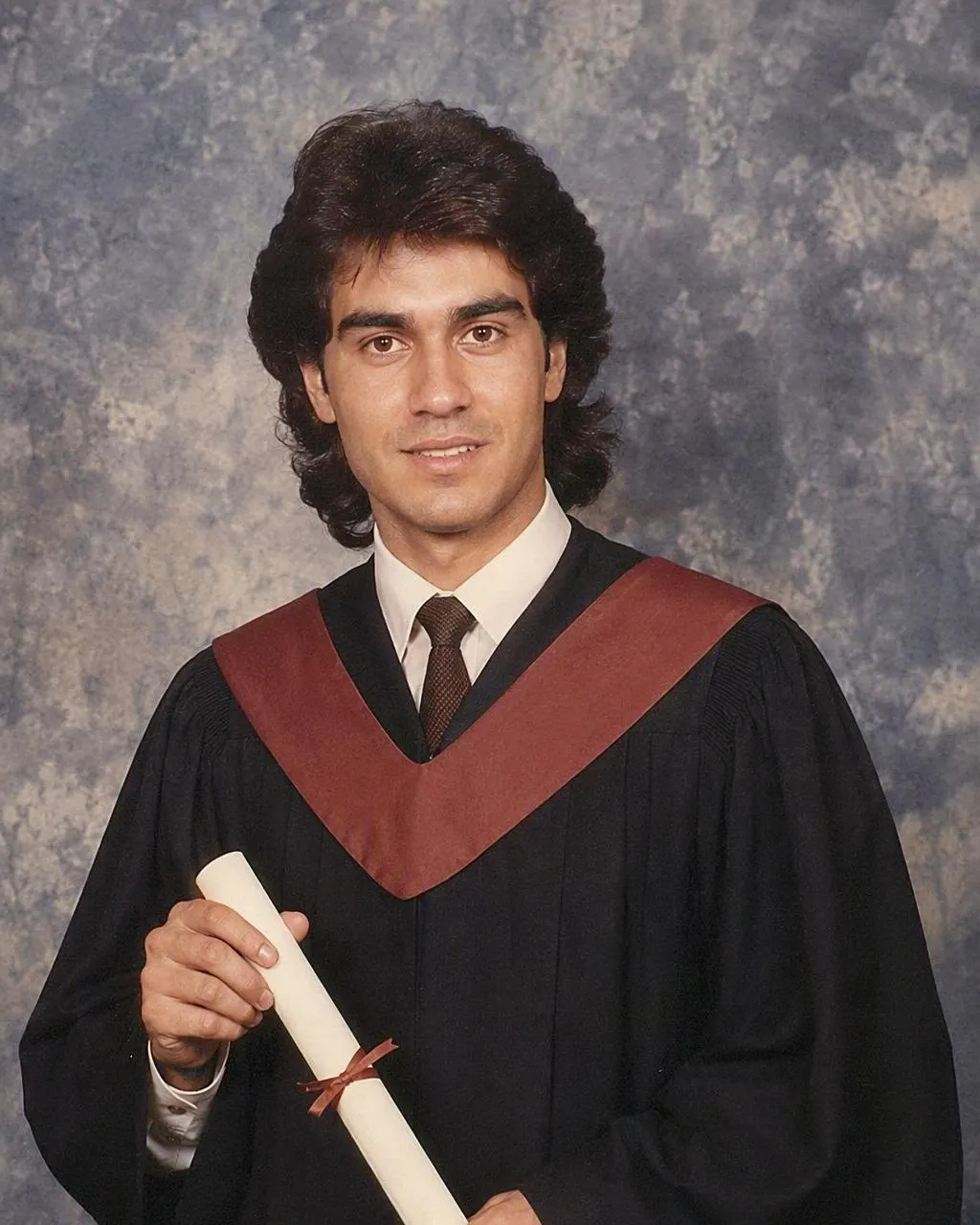 Foto de graduación estilo años 80 generada con IA: fondo de estudio azul jaspeado, luz frontal suave, toga y birrete con peinado ochentero