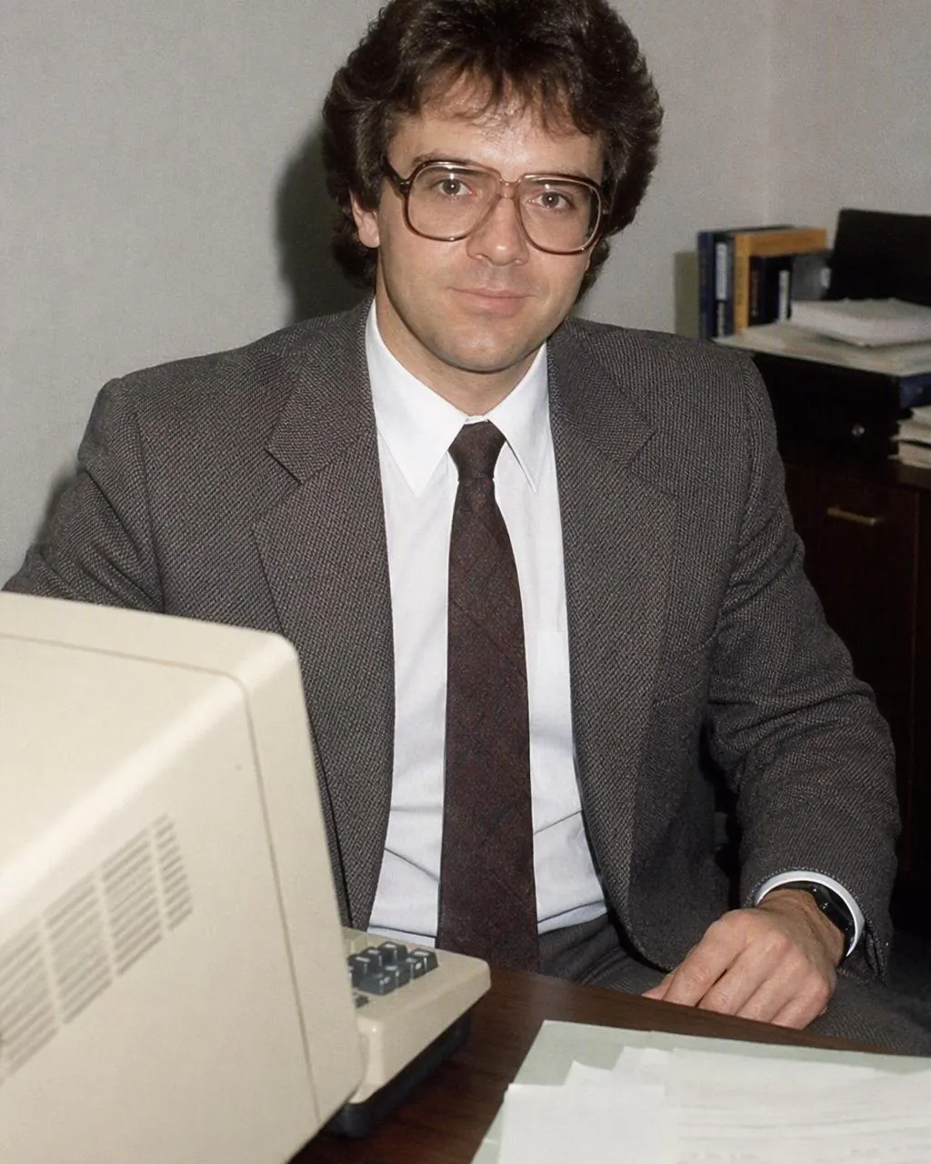 Retrato de oficina con estilo años 80: traje con hombreras, corbata ancha estampada, lentes de armazón grande y pared de madera